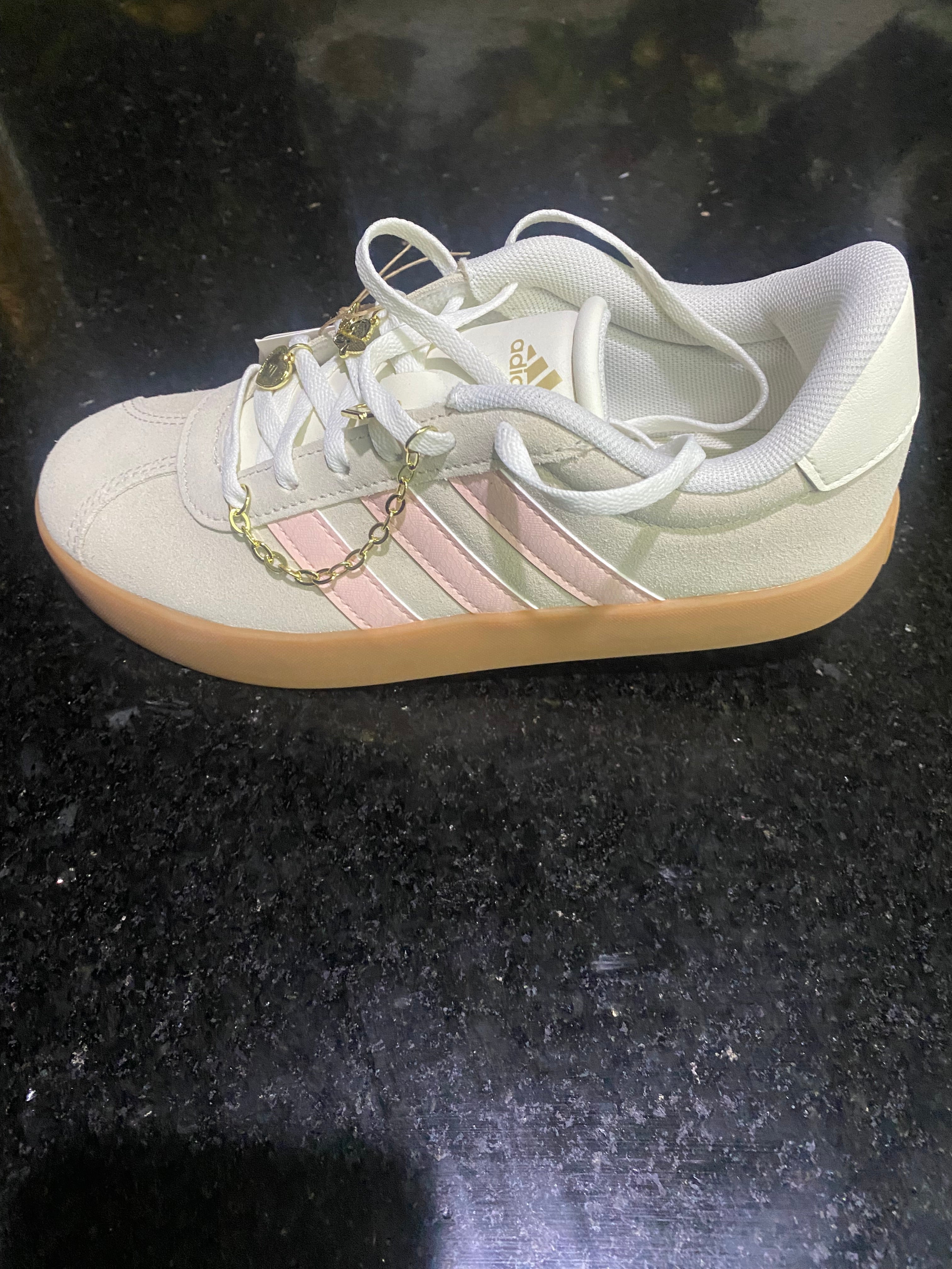 A single Adidas sneaker in off-white with light pink stripes and a gum sole, decorated with a gold chain, shown on a dark speckled countertop.